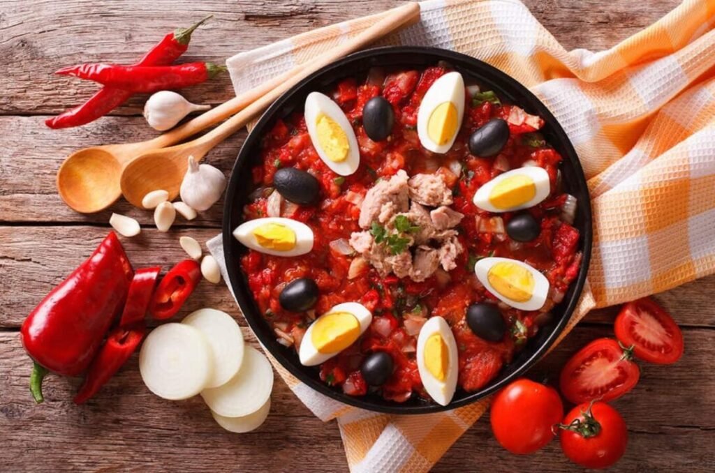 Traditional Mediterranean stew in a black skillet containing tomatoes, red peppers, olives, eggs, tuna, and garnished with herbs, surrounded by ingredients like garlic, onion, and fresh tomatoes on a rustic wooden table.