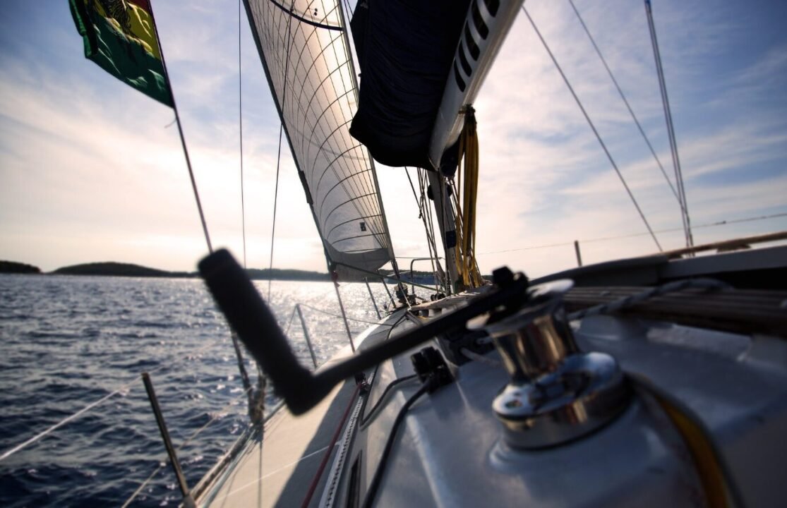 Sailboat deck view at sunset, featuring the mainsail and jib, with a winch in the foreground and a fluttering flag on the left, sailing on tranquil waters.