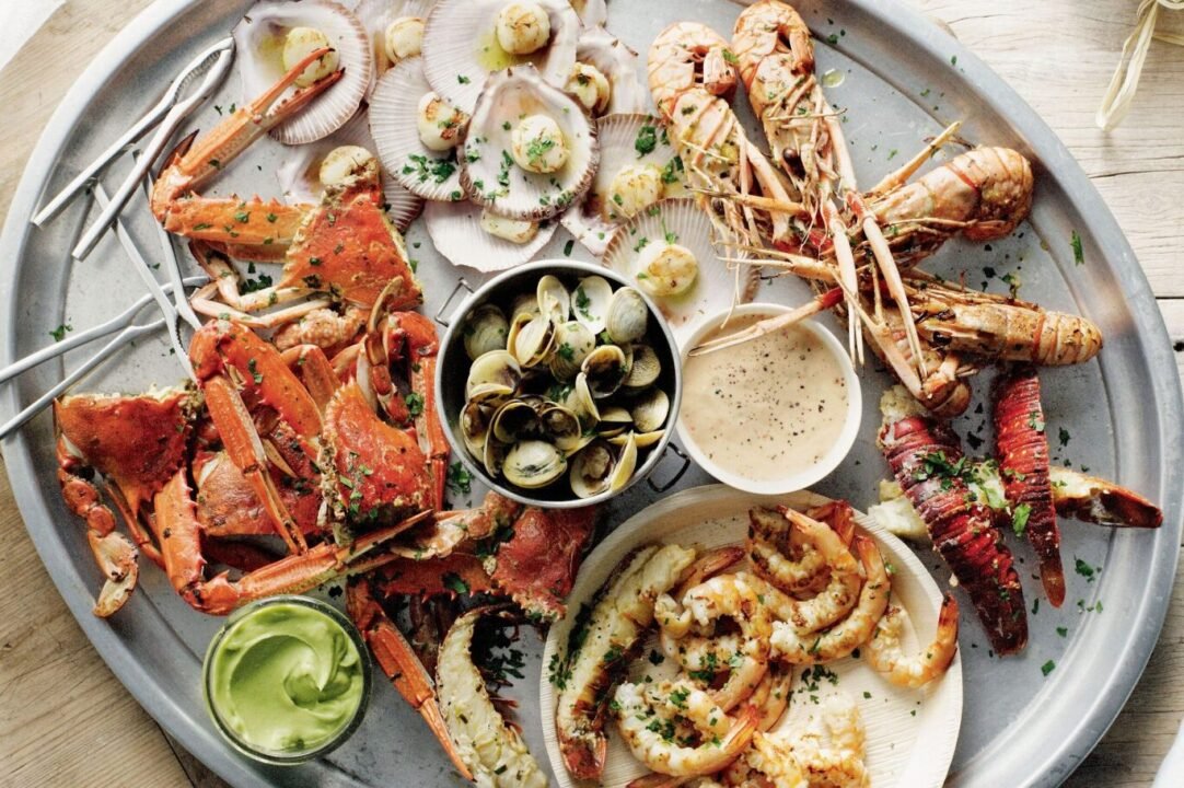Assorted seafood platter featuring crab legs, scallops, clams, prawns, and lobster tails, garnished with herbs and served with dipping sauces on a metal tray.