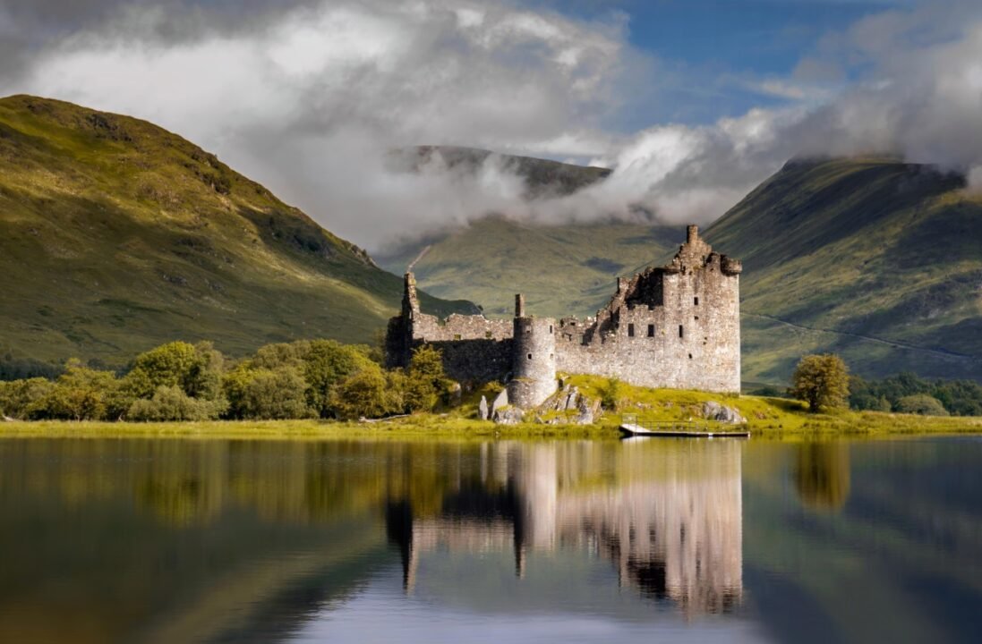 Historic castle on island in calm water with mountain backdrop and cloudy sky.