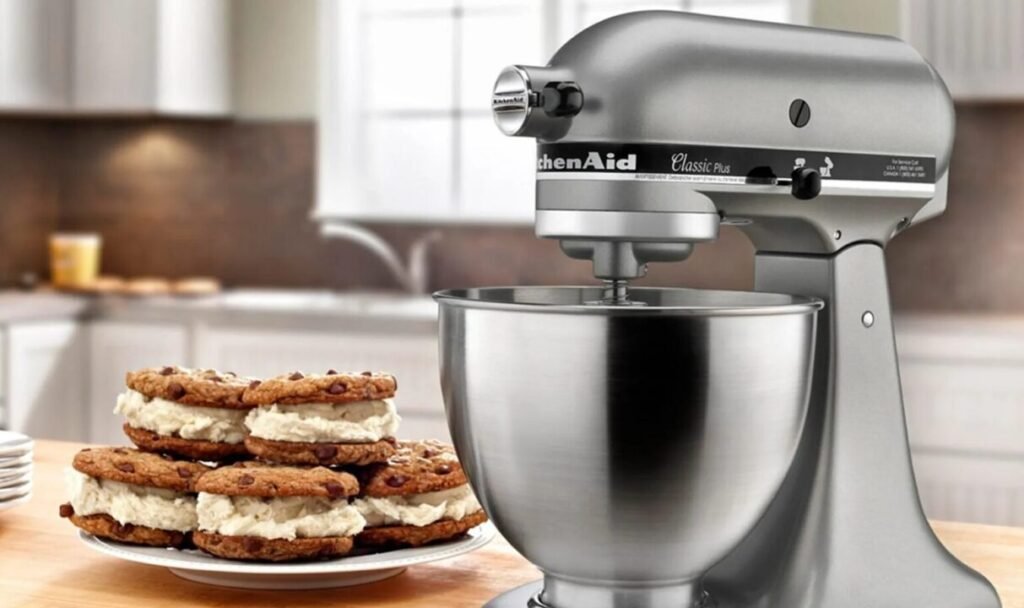 A silver KitchenAid stand mixer on a kitchen counter with a plate of ice cream sandwiches made with chocolate chip cookies in the foreground.