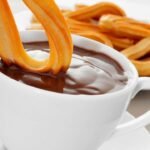 Delicious churros dipped in rich chocolate, with more churros on a plate.