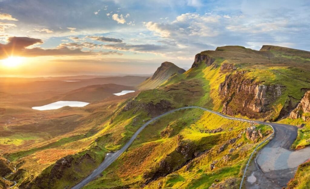 Scenic view of a winding road through lush green hills and rugged cliffs during sunset, with lakes in the distance and dramatic clouds above.