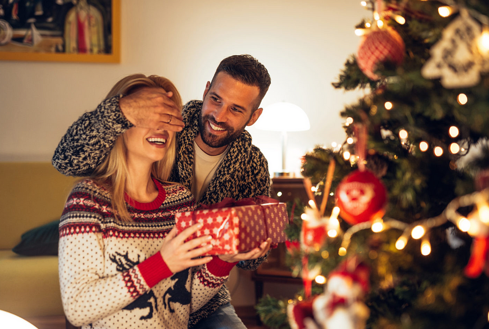 Couple sharing festive gift surprise by Christmas tree, joyful in warmly lit room.