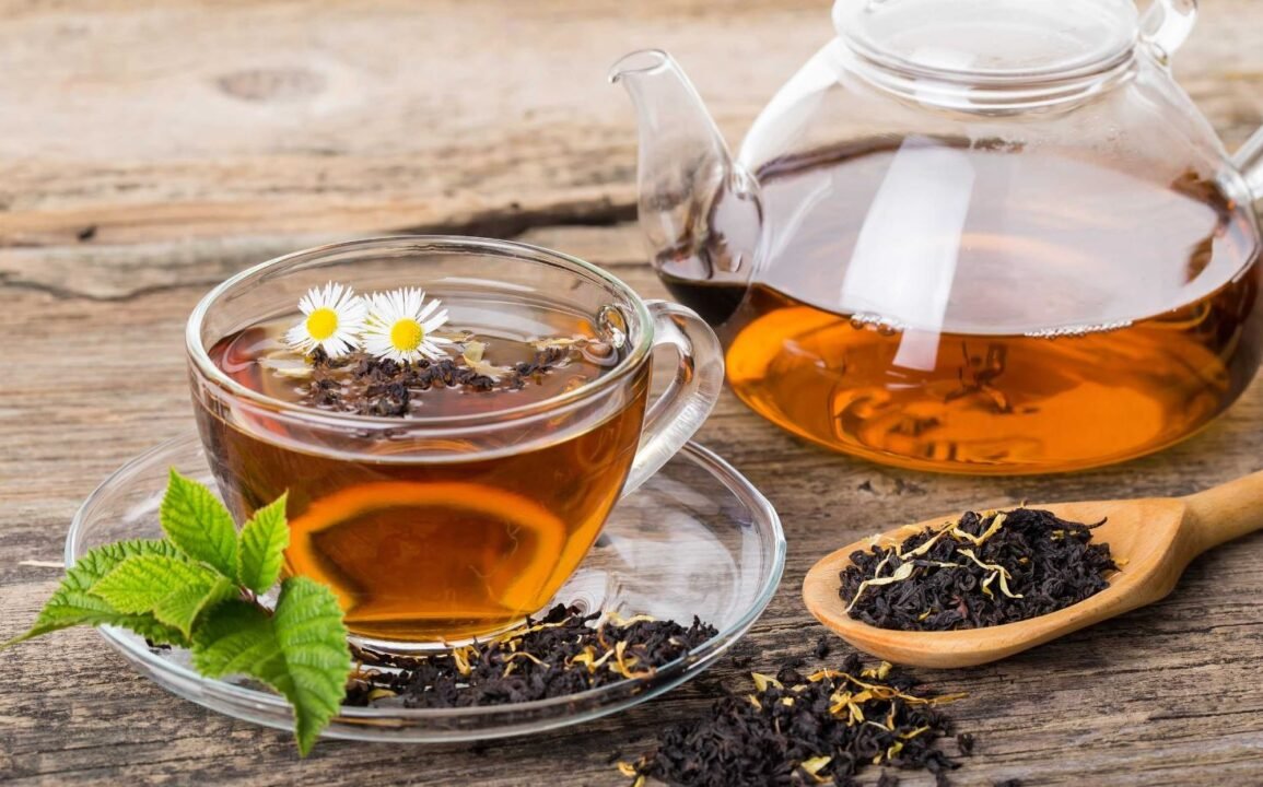 Glass cup of tea with daisies and loose tea leaves on a rustic wooden table, accompanied by a glass teapot and a wooden spoon filled with tea leaves.