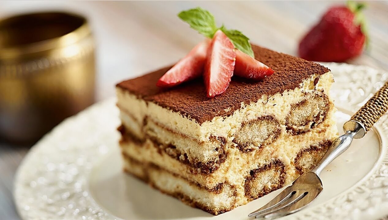 Slice of tiramisu with fresh strawberry slices and mint on top, served on a decorative white plate with a fork, near a blurred metal cup.