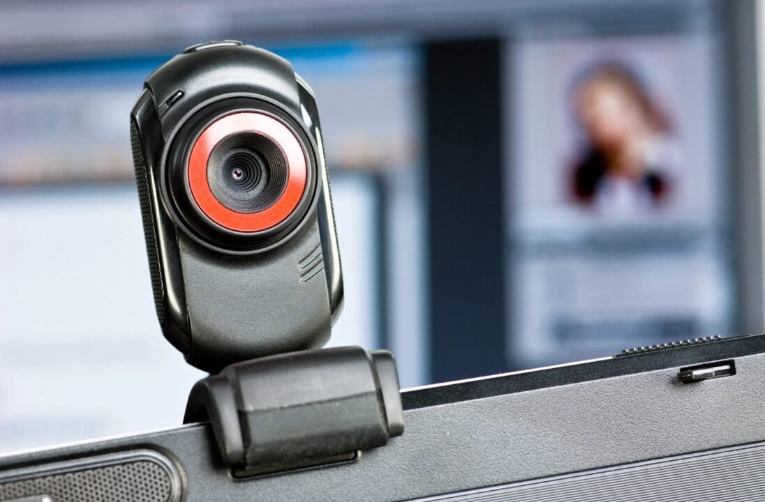 Close-up of a web camera attached to a laptop with a blurred image of a person on the computer screen in the background.