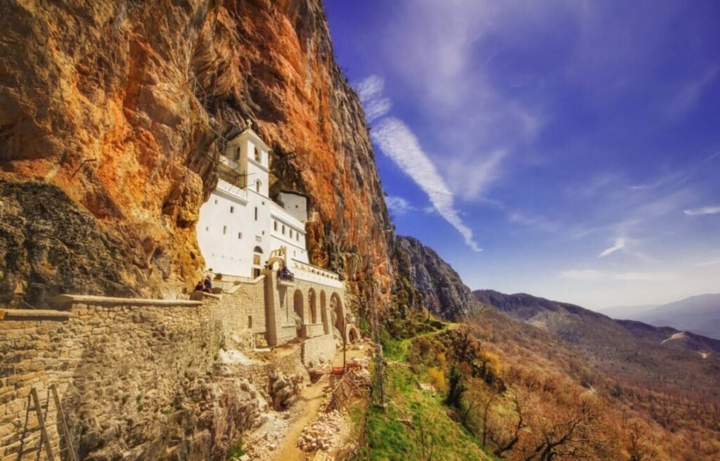 White monastery built into a steep cliff face with a panoramic view of mountains under a blue sky with scattered clouds.