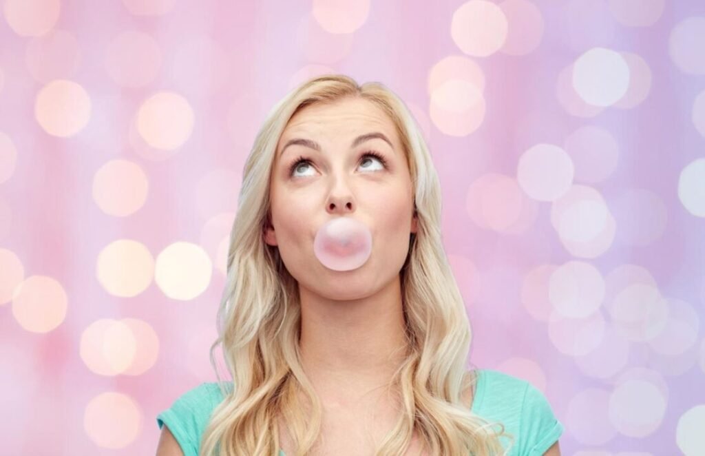 Blonde woman blowing a bubble with gum, looking up, with a pink and purple bokeh background.