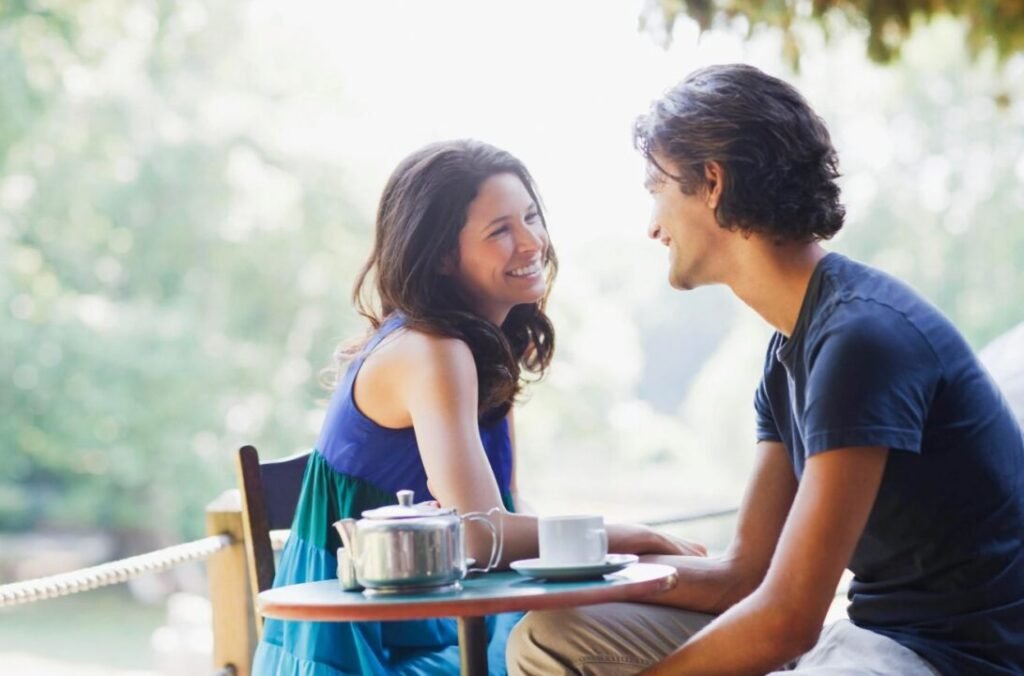 A man and a woman sit at an outdoor café table, smiling and looking at each other. A teapot and two cups are on the table.