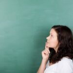 Woman thinking and looking up, standing in front of a green chalkboard.