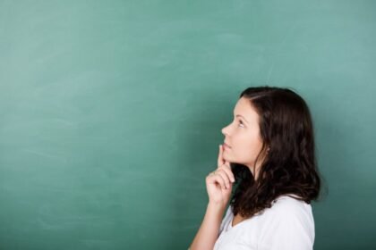 Woman thinking and looking up, standing in front of a green chalkboard.
