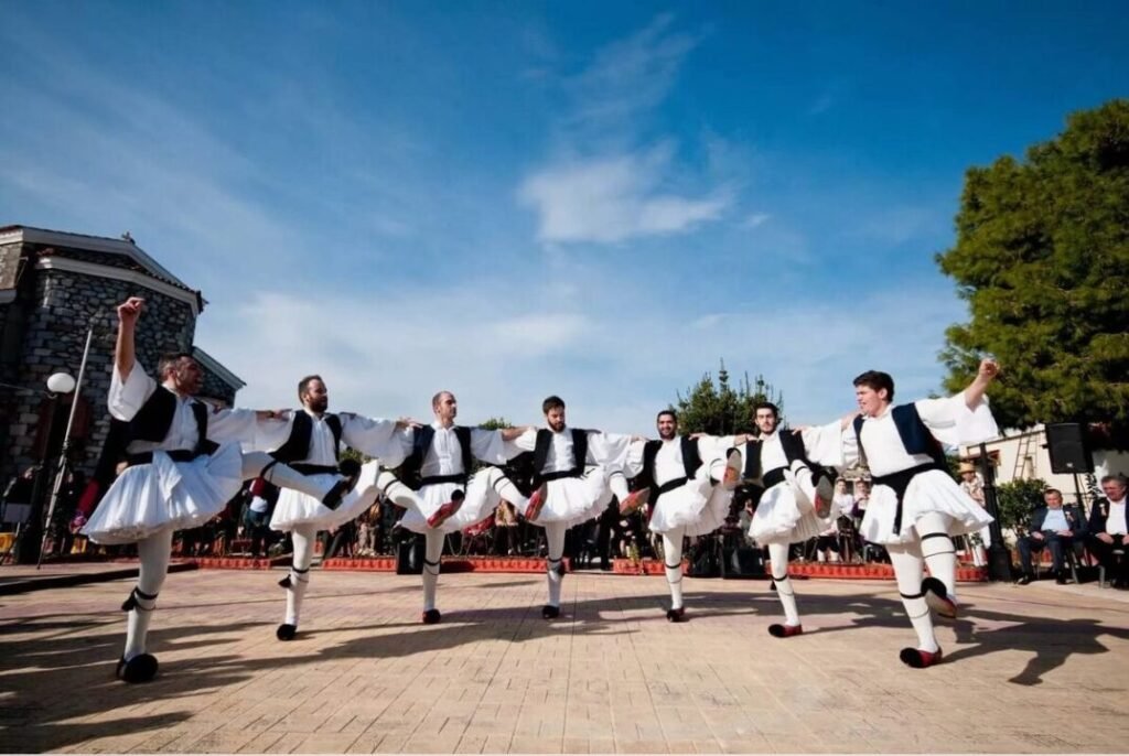 Men in traditional Greek attire performing a synchronized outdoor dance with their legs raised.