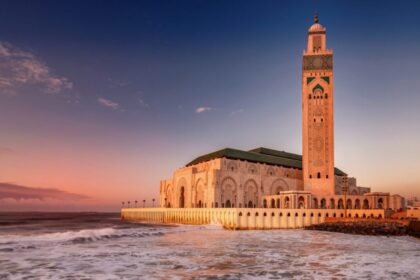A large mosque by the ocean at sunset, featuring intricate architectural details and an ornate minaret under a clear sky with soft pink and purple hues.