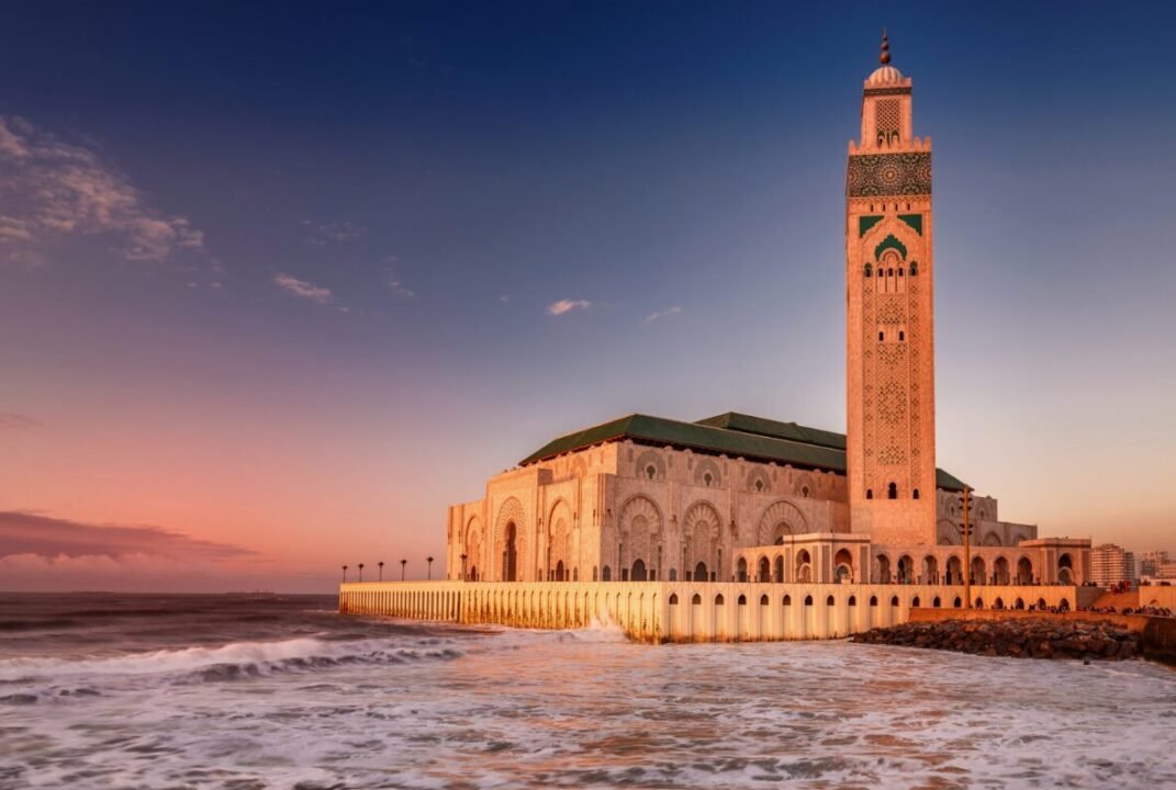 A large mosque by the ocean at sunset, featuring intricate architectural details and an ornate minaret under a clear sky with soft pink and purple hues.