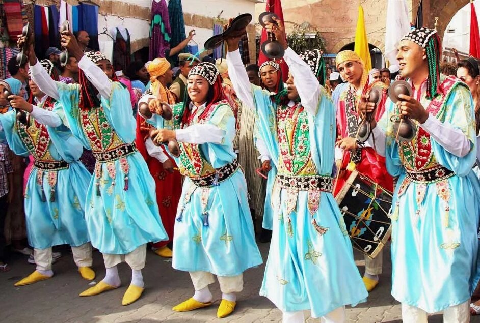 People in brightly colored traditional clothing performing a dance with percussion instruments in a lively outdoor setting with a crowd watching.