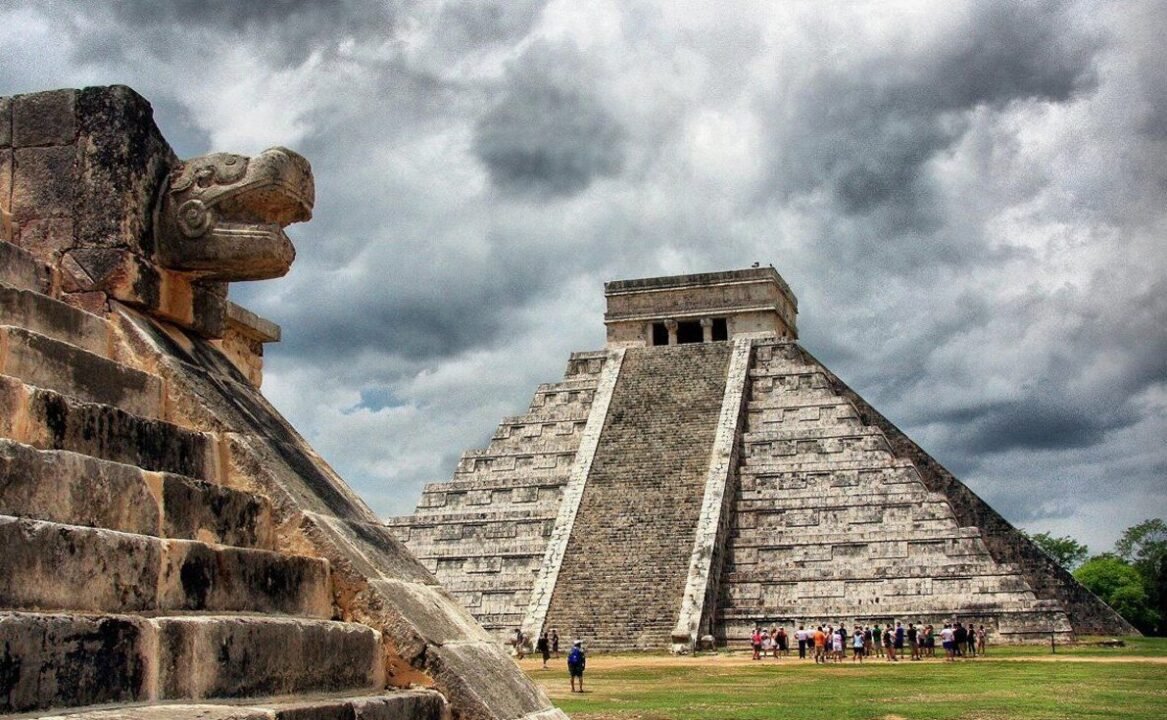 Pyramid of Kukulcán at Chichen Itza with cloudy sky and tourists at the base, featuring a stone carved serpent head on the left.