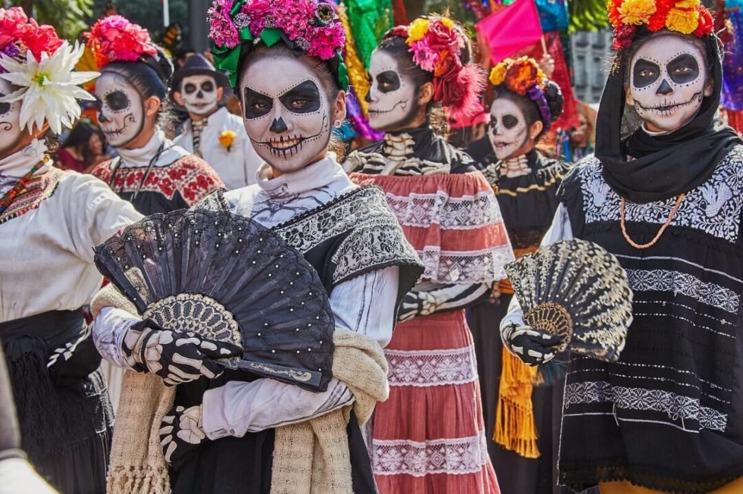 People in traditional costumes and makeup for Dia de los Muertos, holding decorated fans.
