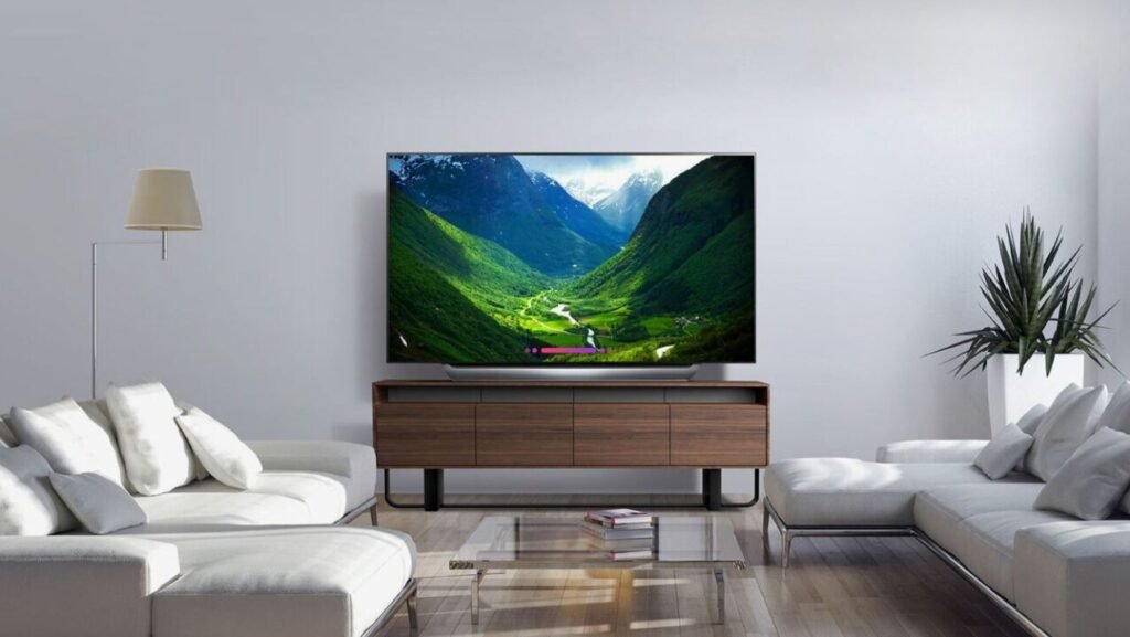 Modern living room with white sofas, a large flat-screen TV displaying a scenic valley, a wooden TV stand, a glass coffee table, a floor lamp, and a potted plant.