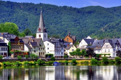 A picturesque village with colorful houses and a prominent church with a tall steeple, situated by a river and surrounded by lush green hills.