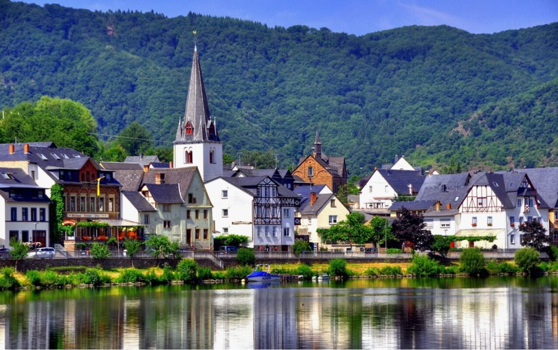 A picturesque village with colorful houses and a prominent church with a tall steeple, situated by a river and surrounded by lush green hills.