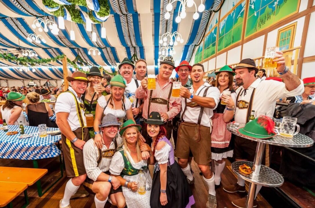 Group of people in traditional Bavarian attire celebrating with beer mugs at a festival inside a large decorated tent.