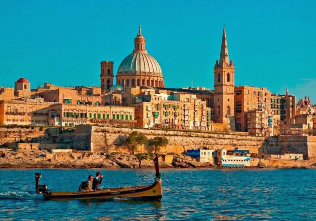 A small boat with two people on the water in front of an old city featuring prominent domed and spired buildings under a clear blue sky.