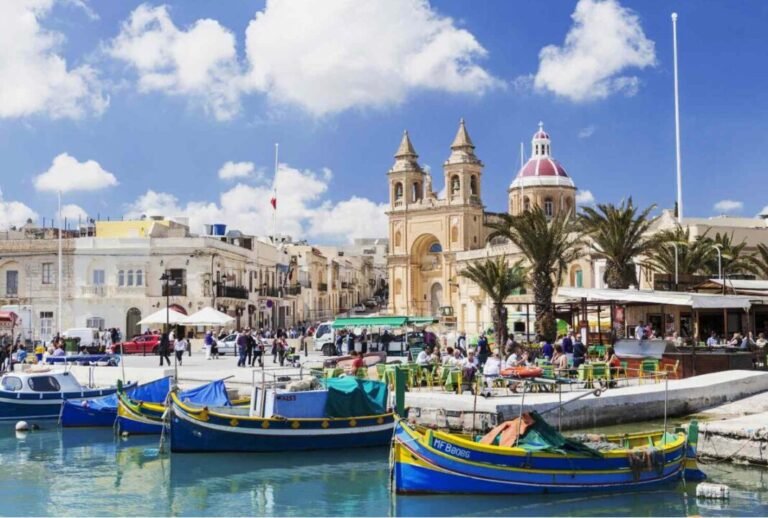 A waterfront plaza with colorful boats docked, people walking and dining outdoors, historic buildings in the background, and a church with twin bell towers and a red dome.