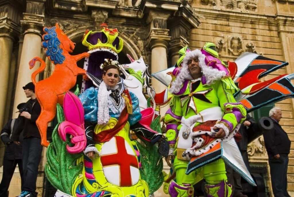 Two people in vibrant, elaborate costumes stand in front of an ornate building facade, with one dressed in a knight and queen-themed outfit and the other in a brightly colored, fantasy-inspired ensemble.