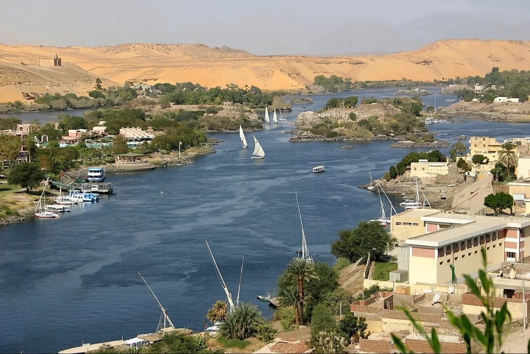 River flowing through a town with boats and sailboats on the water, surrounded by lush greenery and desert hills in the background.