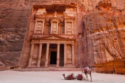 Ancient rock-carved structure with columns and ornate facades, with camels resting in front.