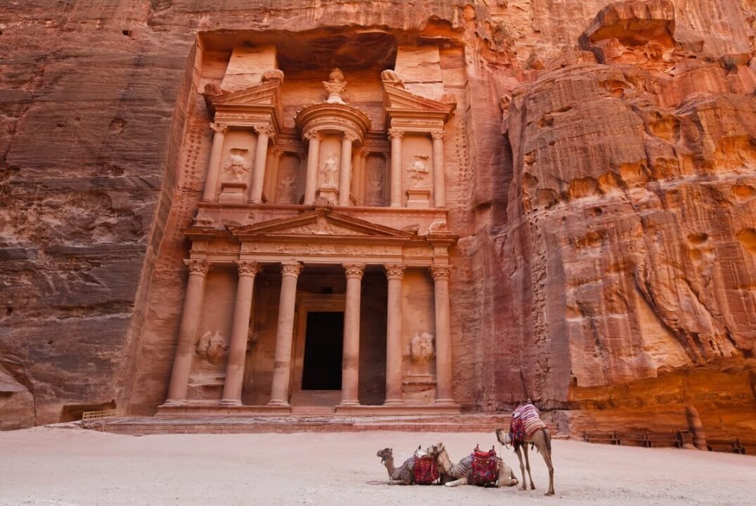 Ancient rock-carved structure with columns and ornate facades, with camels resting in front.