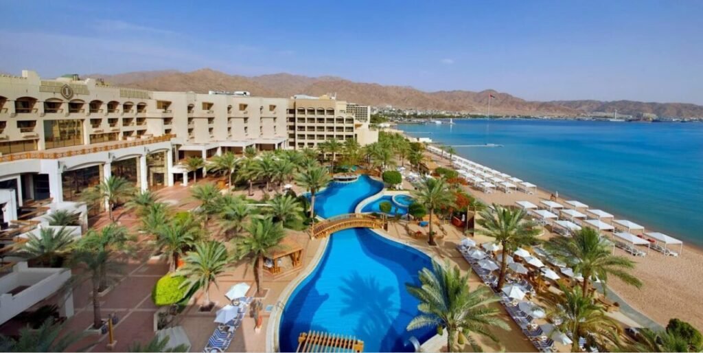 Aerial view of a beachfront resort with a winding swimming pool surrounded by palm trees, lounge chairs, and umbrellas, adjacent to a sandy beach and clear blue sea with mountains in the background.