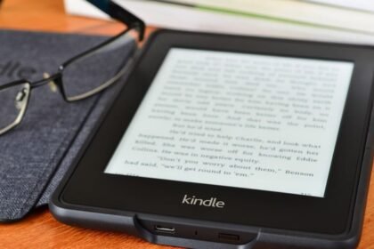 A Kindle e-reader displaying text on a wooden surface next to a pair of glasses.