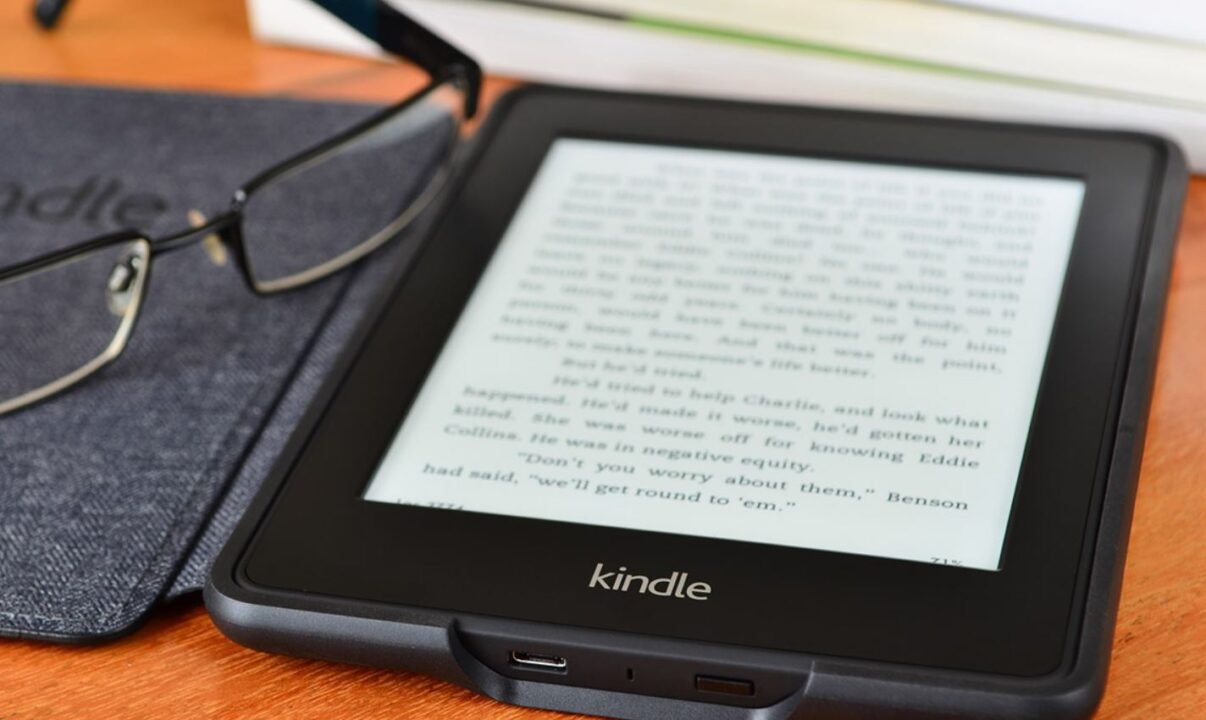 A Kindle e-reader displaying text on a wooden surface next to a pair of glasses.