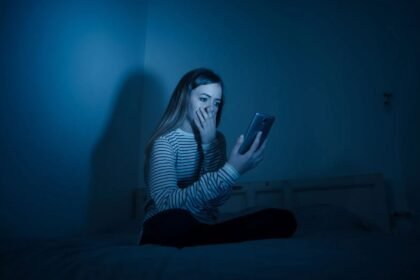 A woman sitting on a bed in a dark room, looking shocked while holding a smartphone, with blue lighting.