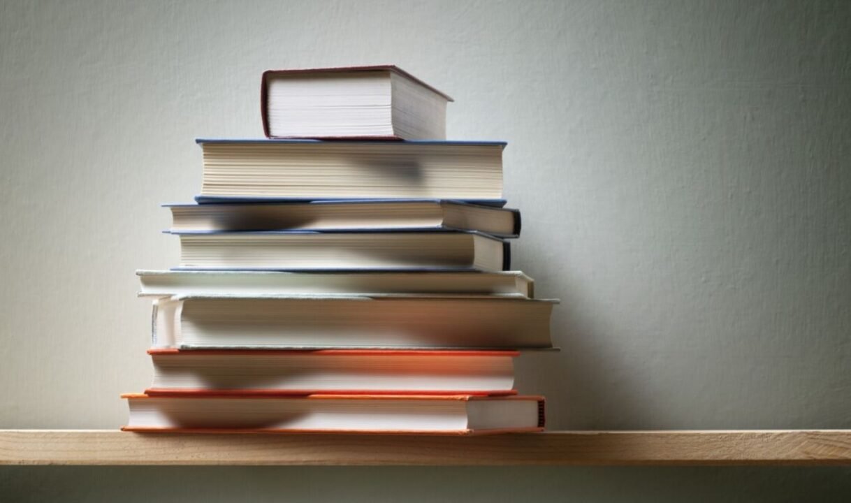 Stack of books on a wooden shelf against a plain wall.