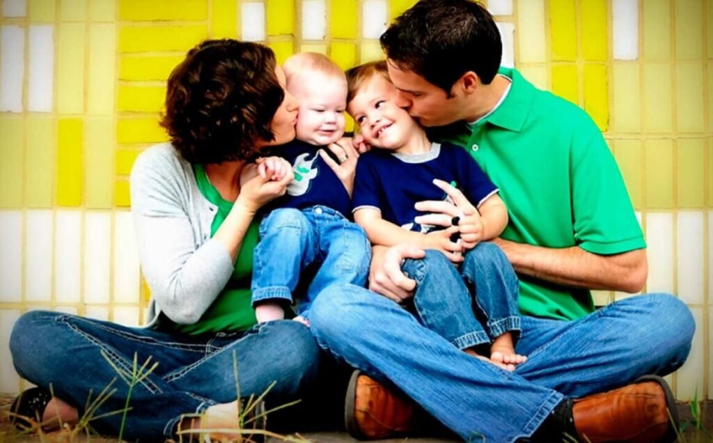 A family of four sitting together, with the parents kissing their two young children, in front of a yellow patterned wall.