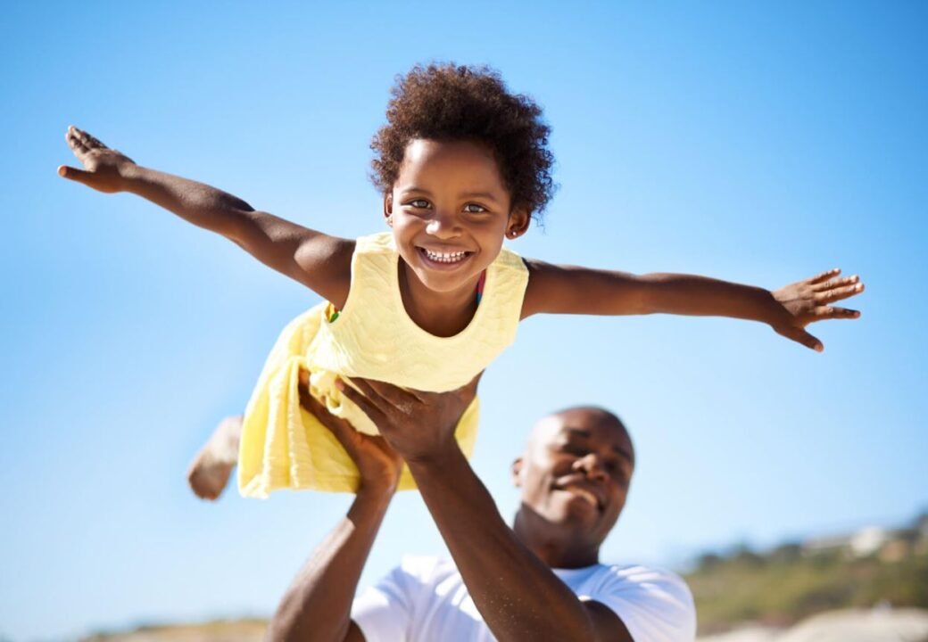 A man playfully lifts a smiling girl in a yellow dress with arms outstretched, against a clear blue sky.