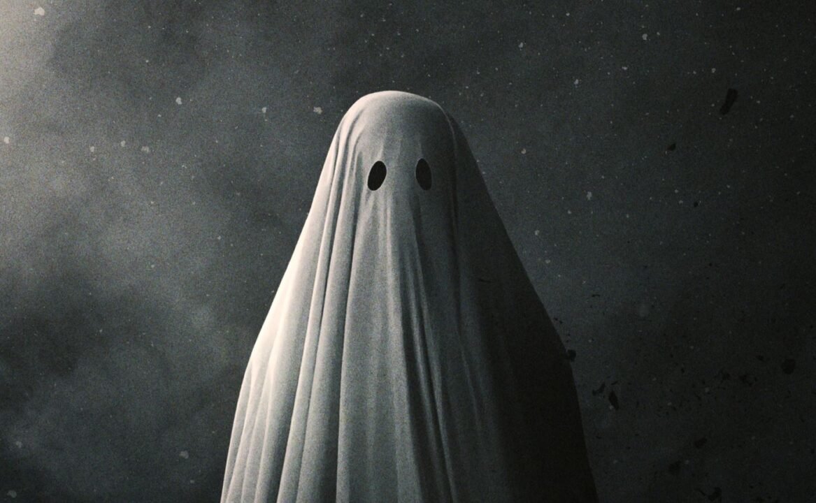 Person dressed as a ghost, covered in a white sheet with two black eye holes, standing against a dark, speckled background.