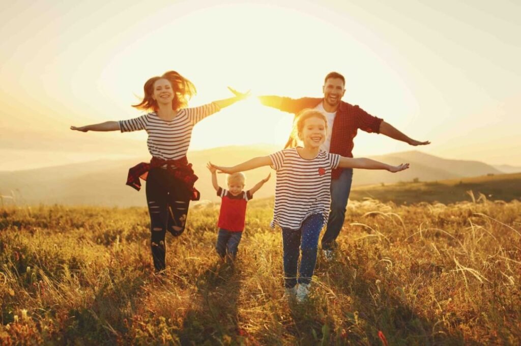 A family of four, with two children and two adults, joyfully running in a grassy field at sunset with their arms outstretched.