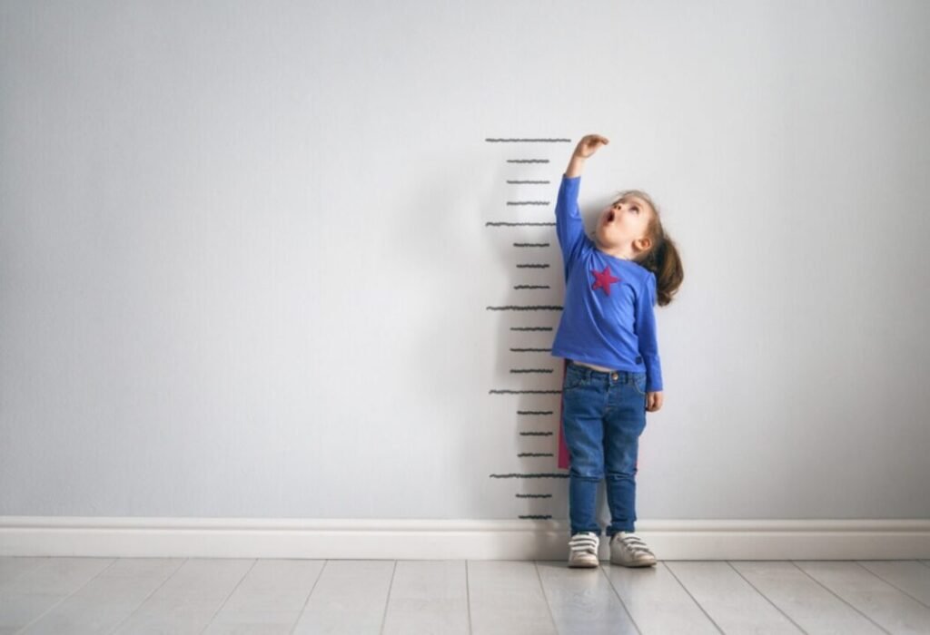 A young child stands next to a height measurement on the wall, reaching up with one hand.