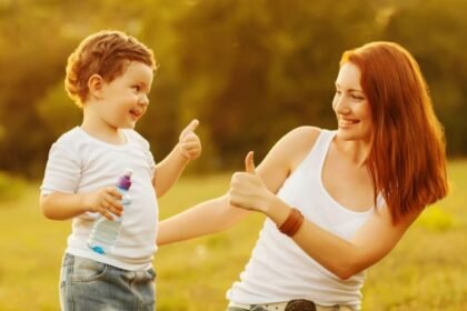 A woman and a child give thumbs up to each other while smiling outdoors.