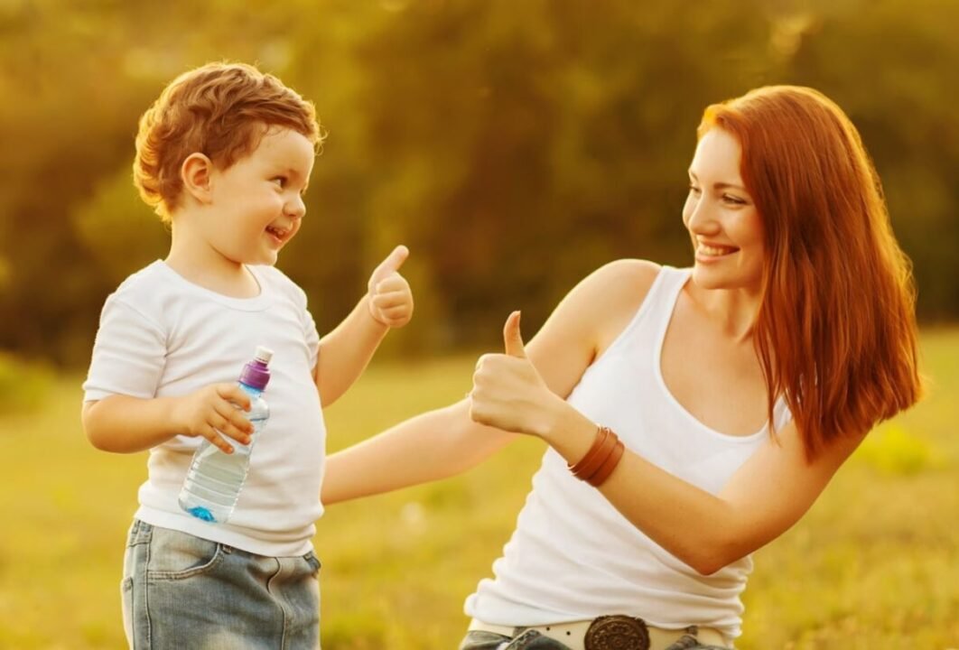 A woman and a child give thumbs up to each other while smiling outdoors.