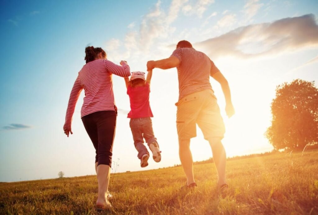 A family walking in a grassy field at sunset, with a child being swung by two adults.