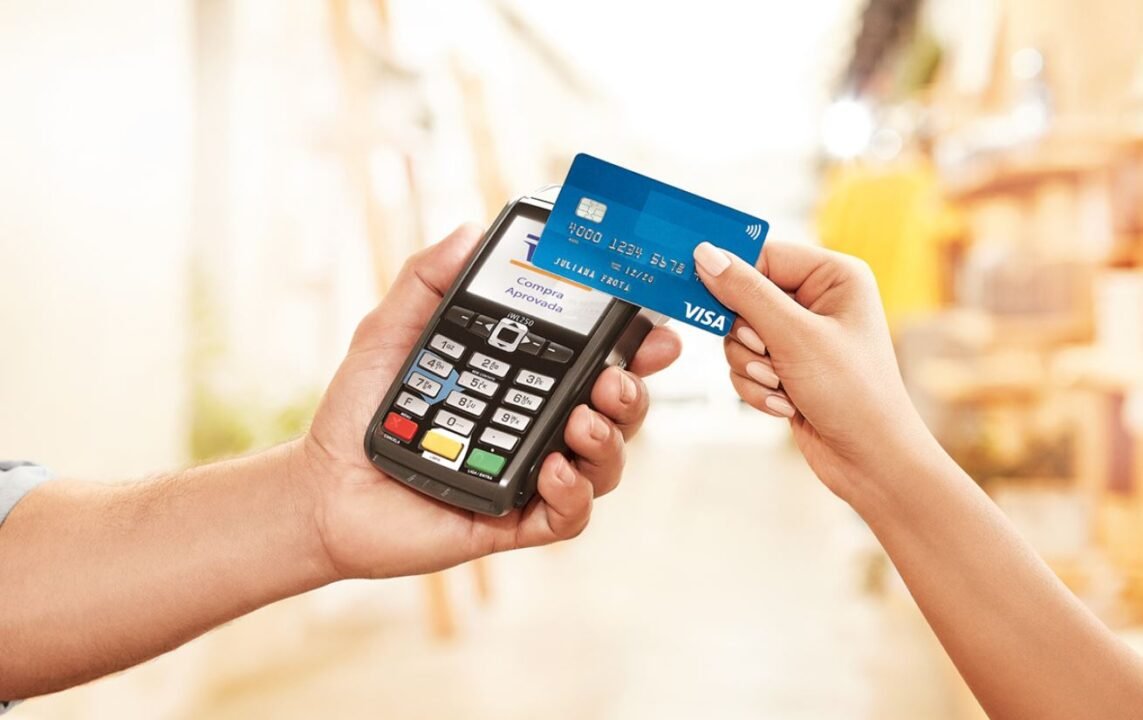 A hand holding a blue contactless Visa card near a card reader for payment.