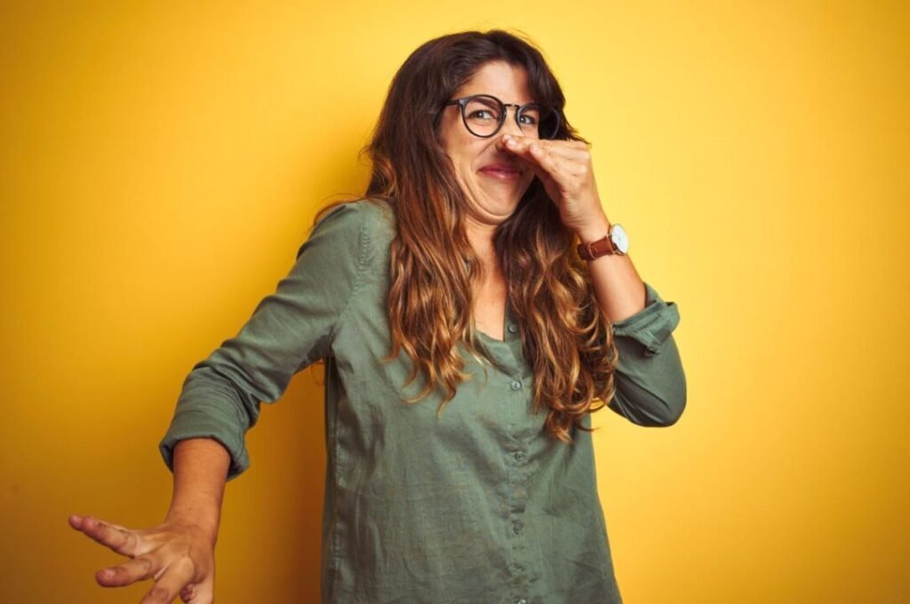 Woman with long hair and glasses holding her nose in disgust against a yellow background.