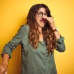 Woman with long hair and glasses holding her nose in disgust against a yellow background.