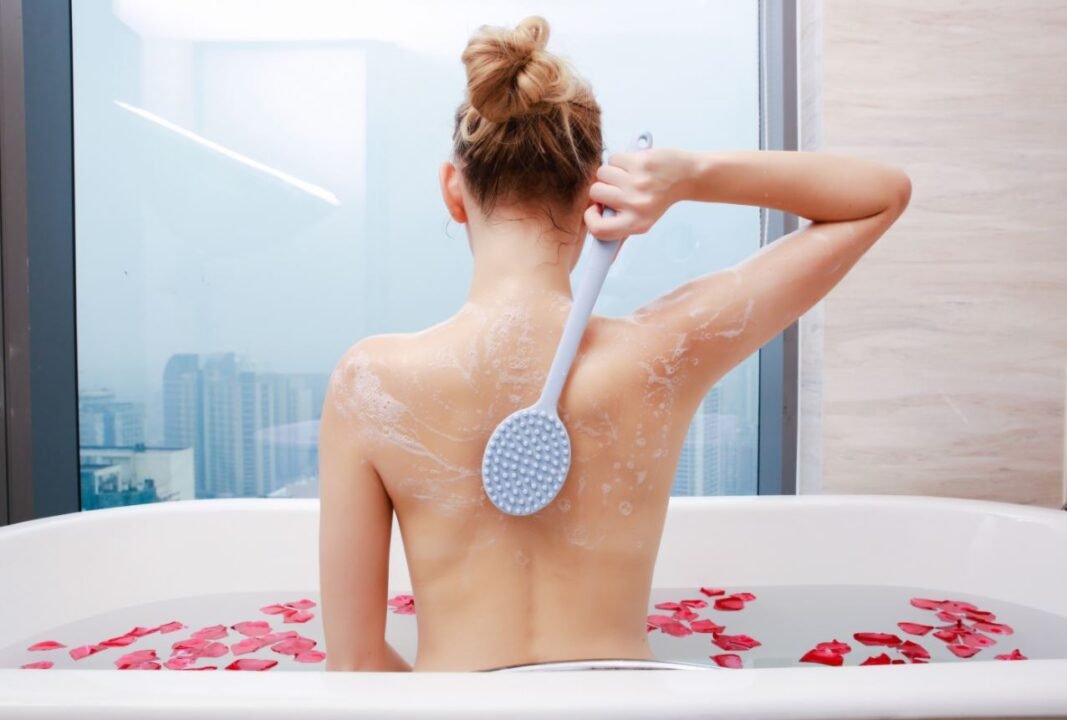 A person in a bathtub with rose petals, using a back scrub brush, next to a window overlooking a cityscape.