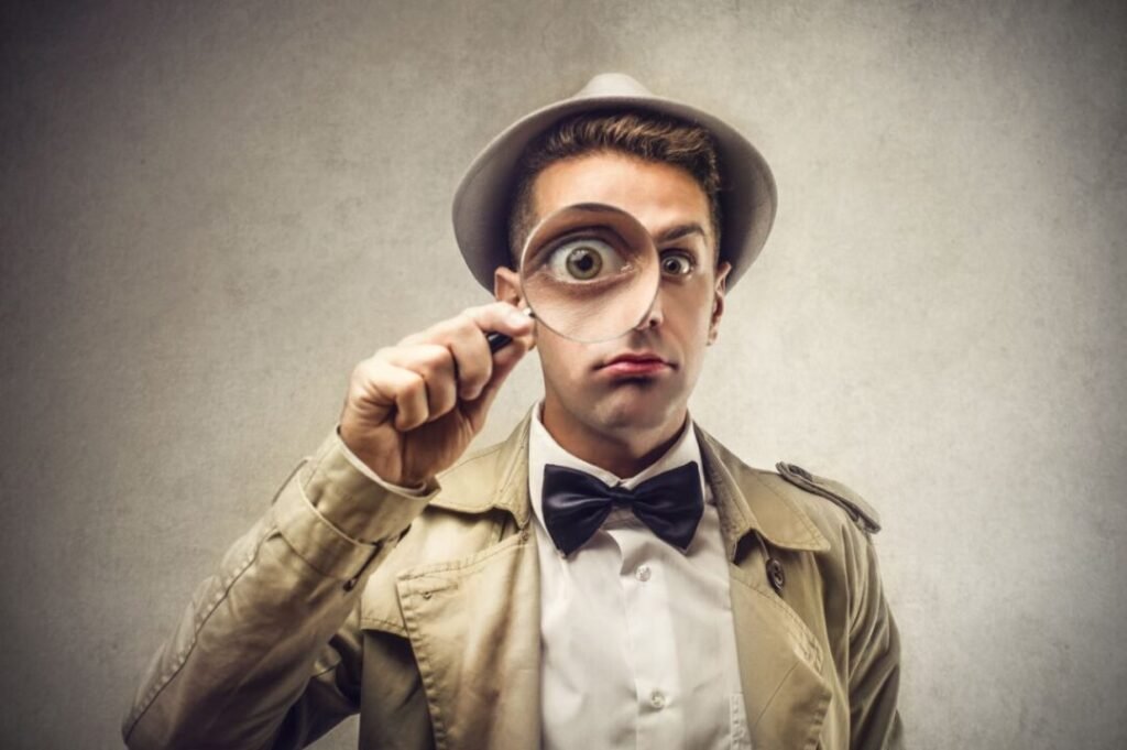 Man in a trench coat and hat holding a magnifying glass to his eye.